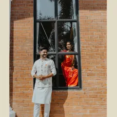 An architectural portrait of an engaged couple, with the groom outside and the bride looking out from a large window.