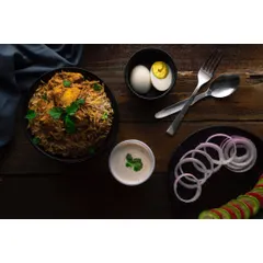 A complete biryani meal composition, shot on a dark wooden surface to create a rustic, traditional feel. The arrangement includes side dishes like raita and boiled egg, showing a full serving suggestion.