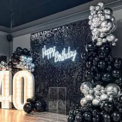 A chic and modern setup for a 40th birthday. The silver sequin wall provides a sparkling backdrop for the neon sign and the large, illuminated marquee numbers, all framed by a black and silver balloon arrangement.