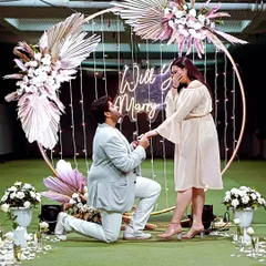 A beautiful moment captured. This modern proposal features a gold ring backdrop decorated with pampas grass and white florals, with delicate fairy lights and a neon sign to complete the romantic, boho-inspired look.