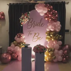 An elegant 'Bride To Be' decoration for a bridal shower. This setup features a custom pink backdrop with gold lettering, framed by a balloon arrangement in shades of pink, rose gold, and chrome gold, with floral accents.