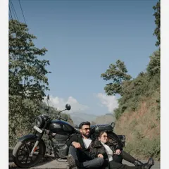 A wide-angle shot of a couple sitting with their motorcycle against a clear blue sky and lush green hills, capturing the freedom of the open road.