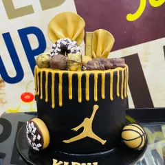 We can design a cake around any theme, like this custom basketball-inspired creation. It features a sleek black fondant finish, a striking gold drip, and is topped with miniature donuts, chocolate bars, and edible basketball details, all centered around the iconic Jordan logo.