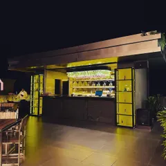 The main bar area of a rooftop cafe, designed to be a focal point. The back-lit shelves and overhead plant installation draw the eye and create a vibrant, modern look.