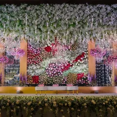 This reception stage is designed to feel like an enchanted forest. A ceiling of hanging white flowers creates a starry night effect, while a dense, multi-colored floral wall provides a breathtaking backdrop for the couple.