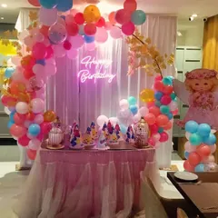 An adorable angel-themed birthday party. The decor features a round balloon arch in pastel colors, a 'Happy Birthday' neon sign, and cute angel cutouts on balloon pillars next to the cake table.