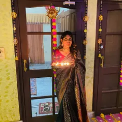 Holding a plate of diyas for Diwali, dressed in a traditional dark silk saree with a contrasting red blouse. This is a classic festive look that is both elegant and timeless.