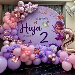 A magical butterfly-themed second birthday for "Hiya," featuring a large circular backdrop in soft purple. The decor is brought to life with a stunning organic balloon garland in shades of pink and purple, accented with metallic and iridescent butterfly cutouts and a large rose gold '2' foil balloon.