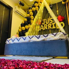 A stunning first-night setup featuring a dramatic rose petal pathway leading to the bed. The decor uses a modern black and gold theme with a balloon arch over a light canopy and "Just Married" foil letters.