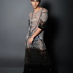A full-length shot of a male model in an avant-garde, deconstructed knit garment. The look is completed with dramatic winged eyeliner and a sleek, wet-look hairstyle.