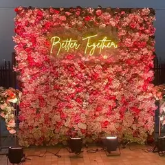 A stunning 'Better Together' neon sign mounted on a dense wall of pink and red flowers. This makes for a perfect photo backdrop at any wedding event.