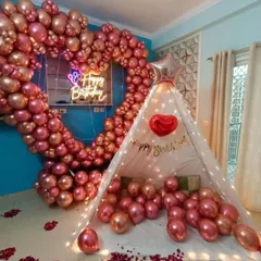 A cozy and romantic birthday surprise featuring a large heart-shaped balloon arch in metallic pink. A small canopy tent and scattered rose petals complete this intimate home setup.