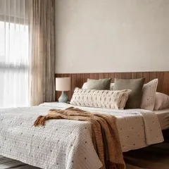 The master bedroom in the Warm Minimal home features lime-washed walls and a wood-paneled headboard. The layered neutral bedding and soft lighting create a deeply restful and elegant space.