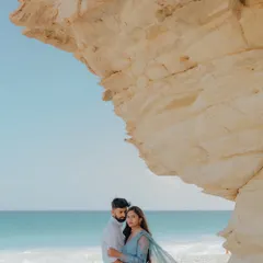 A breathtaking view on the coast of Muscat, Oman. The couple stands beneath a natural rock arch, the turquoise water and blue sky creating a stunning, vibrant backdrop for this serene pre-wedding portrait.