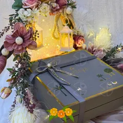 A beautifully packaged gift box for a wedding, featuring a custom grey box with a silver ribbon and delicate floral arrangements. The warm glow of miniature lanterns adds a magical touch to the presentation.