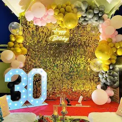 Celebrate a 30th birthday in style with this chic setup. It features a gold sequin wall, an organic balloon garland in pink, black, and gold, and large marquee numbers, creating a perfect spot for party photos.