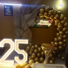 This unique 25th-anniversary setup combines modern and rustic elements. A half-arch of metallic gold balloons is accented with dried palm leaves, framing a "Happy Anniversary" neon sign, with large light-up "25" numbers completing the look.