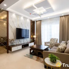 A luxurious living room featuring a floor-to-ceiling marble-finish TV wall with gold inlay details. The design is complemented by a geometric false ceiling and a mix of patterned and solid color seating.