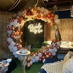 An outdoor terrace setup for a bachelorette party, featuring a circular balloon arch in rose gold and white. The "Bride to be" neon sign glows at the center, creating a perfect photo opportunity next to the jacuzzi.