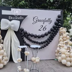 "Gracefully 26." This elegant black and white themed birthday setup is the definition of chic. It features a giant white bow, a string of black pearl-like balloons, and clusters of white balloons with tiny black bow details.