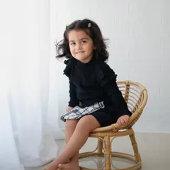 A sweet portrait of a toddler sitting barefoot in a small wicker chair by a window, looking at the camera.
