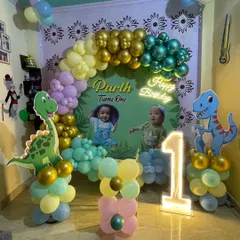 A complete dinosaur theme for a first birthday party. This decoration includes a personalized backdrop, character cutouts of a T-Rex and Brontosaurus, a multi-colored balloon arch, and a light-up number '1' to mark the special milestone.