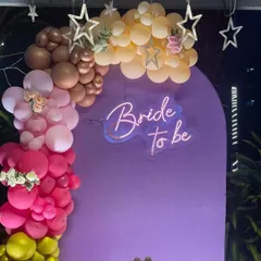 A vibrant and colorful 'Bride to be' setup. This design uses a purple arch backdrop with a neon sign, framed by a multi-colored balloon garland in shades of pink, yellow, and nude, perfect for a fun bachelorette party.