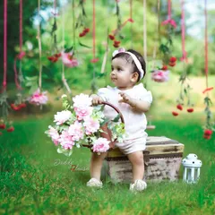 An outdoor sitter session in a beautiful green park. This little girl is sitting on a vintage-style trunk holding a basket of pink flowers, with a lovely floral backdrop hanging from the trees.
