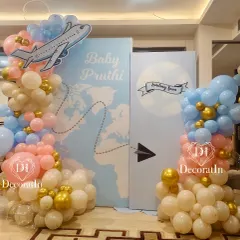 "Baby Pruthi is landing soon." This creative travel-themed baby shower decor features a world map backdrop, an airplane cutout, and a cloud of pink, blue, and gold balloons to celebrate the upcoming arrival.