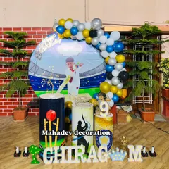 A creative cricket theme first birthday party for a young fan. This outdoor setup includes a custom backdrop showing a stadium, themed pedestals with cricket motifs, and a blue, yellow, and silver balloon garland.
