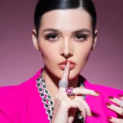 A powerful and intriguing shot from a high-fashion editorial. The styling combines a bold pink blazer with a statement multi-gemstone necklace and vibrant rings, creating a look that is both playful and commanding.