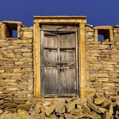 Set against a brilliant blue sky, this rustic wooden door is part of a crumbling stone structure in Kuldhara, Rajasthan. The scattered stones in the foreground add to the feeling of a place forgotten by time.