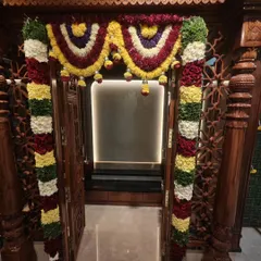 A close-up of the intricate floral garlands decorating the entrance of a traditional wooden pooja room for a housewarming ceremony. We use a mix of colorful flowers for a festive look.