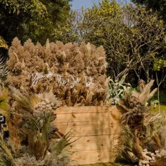 We traded classic florals for hay, pampas, and dried botanicals to create this bold, earthy aesthetic. The "You're The One" neon sign adds a modern, romantic touch to the sophisticated barnyard-chic setup.