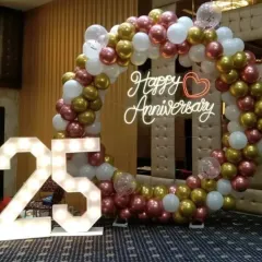 Celebrating a 25th anniversary with style. This decor includes a circular balloon arch in rose gold, gold, and white, a 'Happy Anniversary' neon sign, and large marquee numbers, perfect for creating a memorable photo opportunity in a party hall.