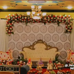 A traditional stage setup combining a patterned fabric backdrop with a heavy floral garland of red and white roses. The decor includes cow figurines, brass lamps, and flower vases, elements often used in auspicious ceremonies like housewarmings or baby showers.