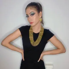 A full-length shot of a glamour fashion look. The makeup features a dramatic blue and green winged eyeshadow, complemented by a bold gold necklace and a sleek high ponytail for a modern, Cleopatra-inspired vibe.