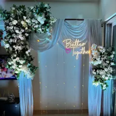 An elegant 'Better Together' backdrop perfect for an engagement or anniversary party. We use white drapes, fairy lights, a neon sign, and lush white artificial flower arrangements.