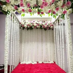 A four-poster mandap design with a canopy of pink and white flowers. The posts are draped with strings of white flowers, creating a beautiful and full look over a red velvet base.