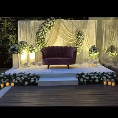 A chic and modern stage for a ring ceremony. The design features a blush pink sofa, a backdrop of white drapes, and a semi-circular floral arch, all illuminated by soft candlelight.