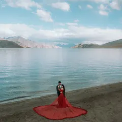 An aerial view capturing the grandeur of Ladakh, with the couple standing by the crystal-clear lake. Her flowing red dress creates a stunning contrast against the blue water.