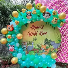 A 'Wild One' first birthday party with a jungle theme. This setup uses a custom printed backdrop and a balloon arch with green and gold chrome balloons and animal character foils.