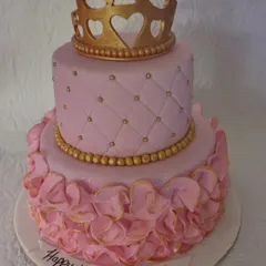 An elegant two-tiered princess cake in pink and gold. The top tier has a quilted pattern, while the bottom is covered in delicate ruffles, all topped with a golden crown.