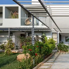 A view of the shaded walkway and private sit-outs at the SAMAM House. A metal pergola structure supports climbing plants, creating a green, living canopy that cools the space and enhances the connection to the garden.