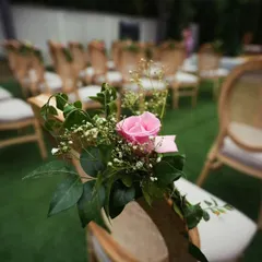 A close-up of a simple yet beautiful chair decoration for an outdoor ceremony. A single pink rose with baby's breath adds a touch of elegance to the rustic wooden chairs.