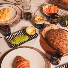 A complete gourmet breakfast spread to inspire your mornings. This table features scrambled eggs, sliced avocado, fresh bread, a croissant, and a variety of our pantry jars like seasonal jam, basil pesto, and truffle hummus.