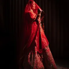 A dramatic bridal portrait with the bride standing in near darkness, her form illuminated by a single light source. The red of her lehenga and the silhouette of her veil create a powerful and artistic image.