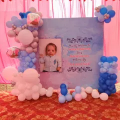A personalized naming ceremony decoration featuring a custom backdrop with the baby's photo and event details. The setup is framed with balloon clusters in shades of pink, blue, and white.