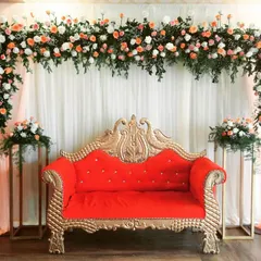 An elegant and simple stage design for an engagement or reception. The backdrop of white drapes is accented with a beautiful floral arch, and a classic red and gold sofa provides seating for the main guests.