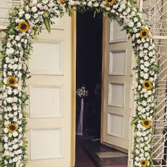 A classic and cheerful floral archway for a church wedding entrance. The arch is covered in white chrysanthemums and bright yellow sunflowers, creating a welcoming and joyous first impression.